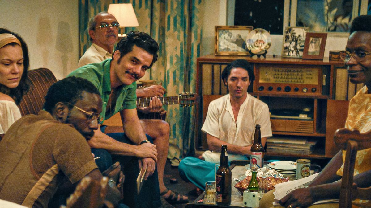 A group of people sitting down and talking. A man in a green shirt is smiling and looking at the camera.